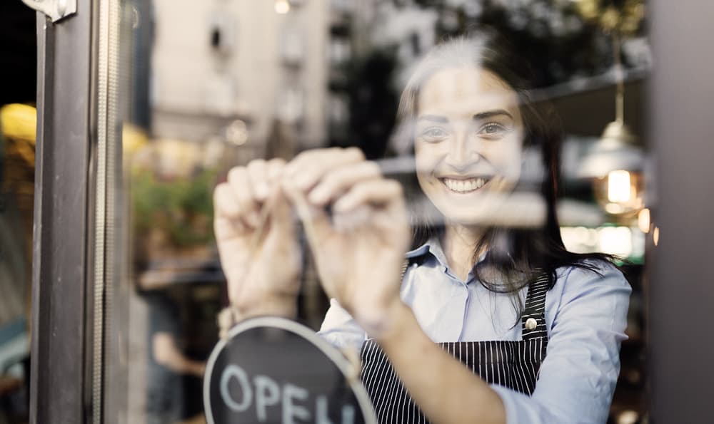 Eine lächelnde Frau in einer gestreiften Schürze öffnet ein Café und richtet ein Schild an der Glastür aus. Die Szene ist warm und einladend, mit verschwommener Straßenansicht draußen.
