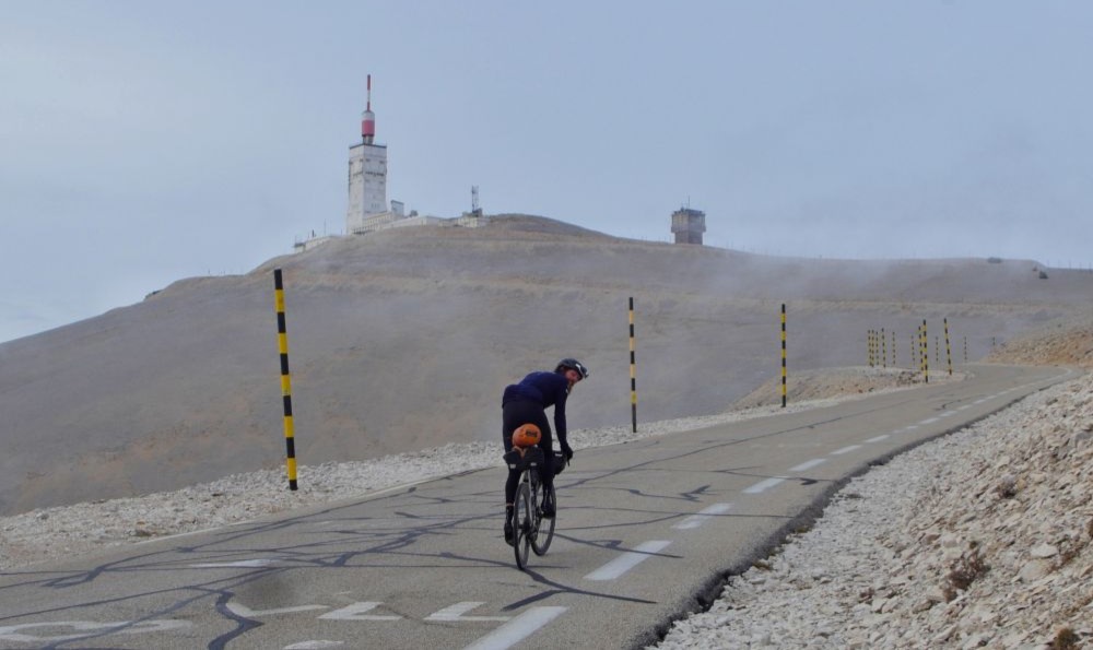 Man sieht den Extremsportler Jonas Deichmann auf einem Rennrad einen Berg in einer sehr kargen Landschaft auf einer Straße hinauffahren. Er dreht sich beim Fahren um und schaut in die Kamera. Oben am Berg steht eine Art Leuchtturm.