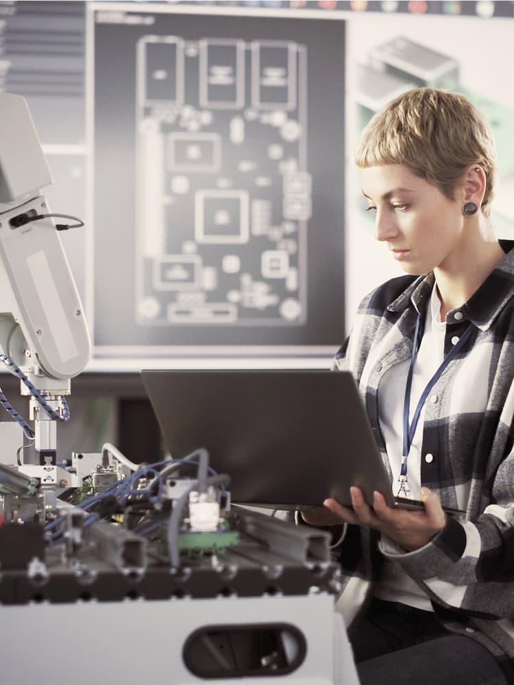 Eine junge blonde Frau mit kurzen Haaren sitzt vor einer Werkbank mit elektronischen Geräten und einem Roboterarm. Sie hält einen Laptop in der Hand über ihrem Schoß, in den sie hineinschaut. Im Hintergrund sieht man verschwommen einen großen Bildschirm mit technischen Zeichnungen.