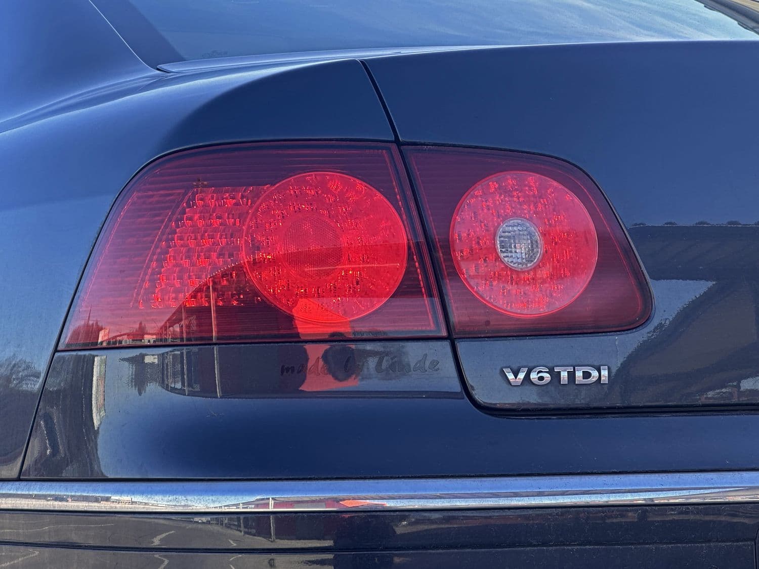 Nahaufnahme des Hecks eines schwarzen Autos mit roten Rückleuchten und einer verchromten V6TDI-Plakette, die das Sonnenlicht reflektiert. Das Bild vermittelt einen eleganten und modernen Eindruck.