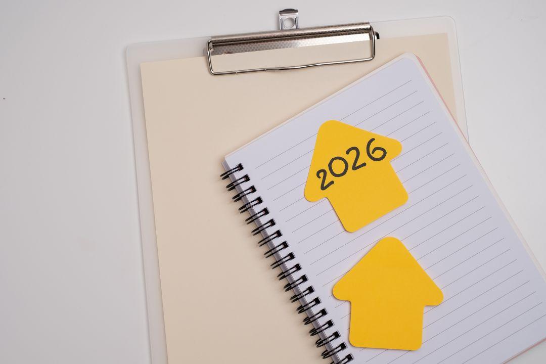 Spiralblock mit zwei gelben, pfeilförmigen Haftnotizen, eine davon mit der Aufschrift „2026“, auf einer linierten Seite. Unter dem Notizbuch befindet sich ein Klemmbrett.