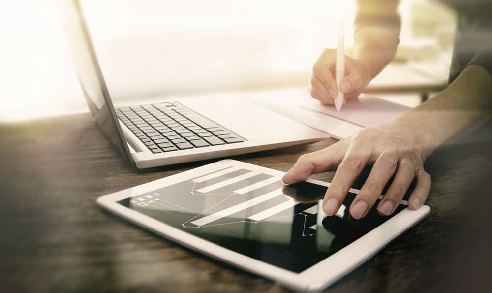 Das Bild zeigt eine Person, die an einem Schreibtisch mit einem Laptop, einem Tablet und einem Notizblock arbeitet. Die Person benutzt einen Stift, um auf den Notizblock zu schreiben, während sie gleichzeitig mit dem Tablet interagiert, das ein Balkendiagramm anzeigt