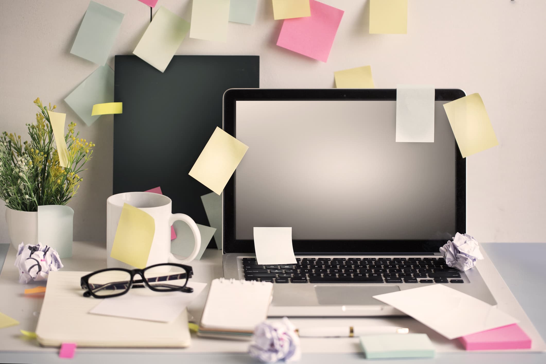 Ein unordentlicher Arbeitsplatz mit einem Laptop, vielen bunten Haftnotizen, einer Tasse, einer Topfpflanze, zerknülltem Papier, einer Brille und einem Notizbuch.