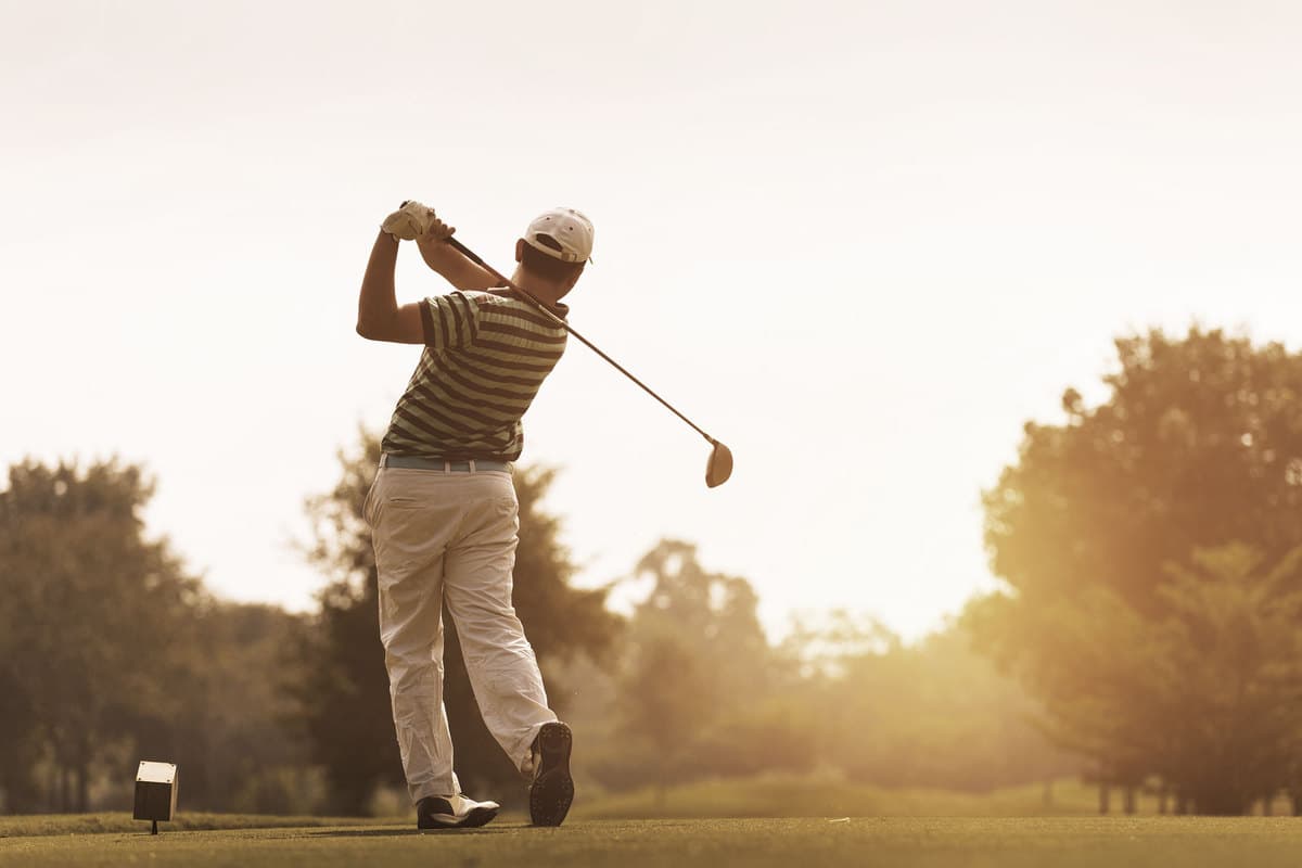 Eine Person beim Golfschwung, mit gestreiftem Shirt, weißer Hose und weißer Kappe. Im Hintergrund Bäume und Sonnenuntergang.