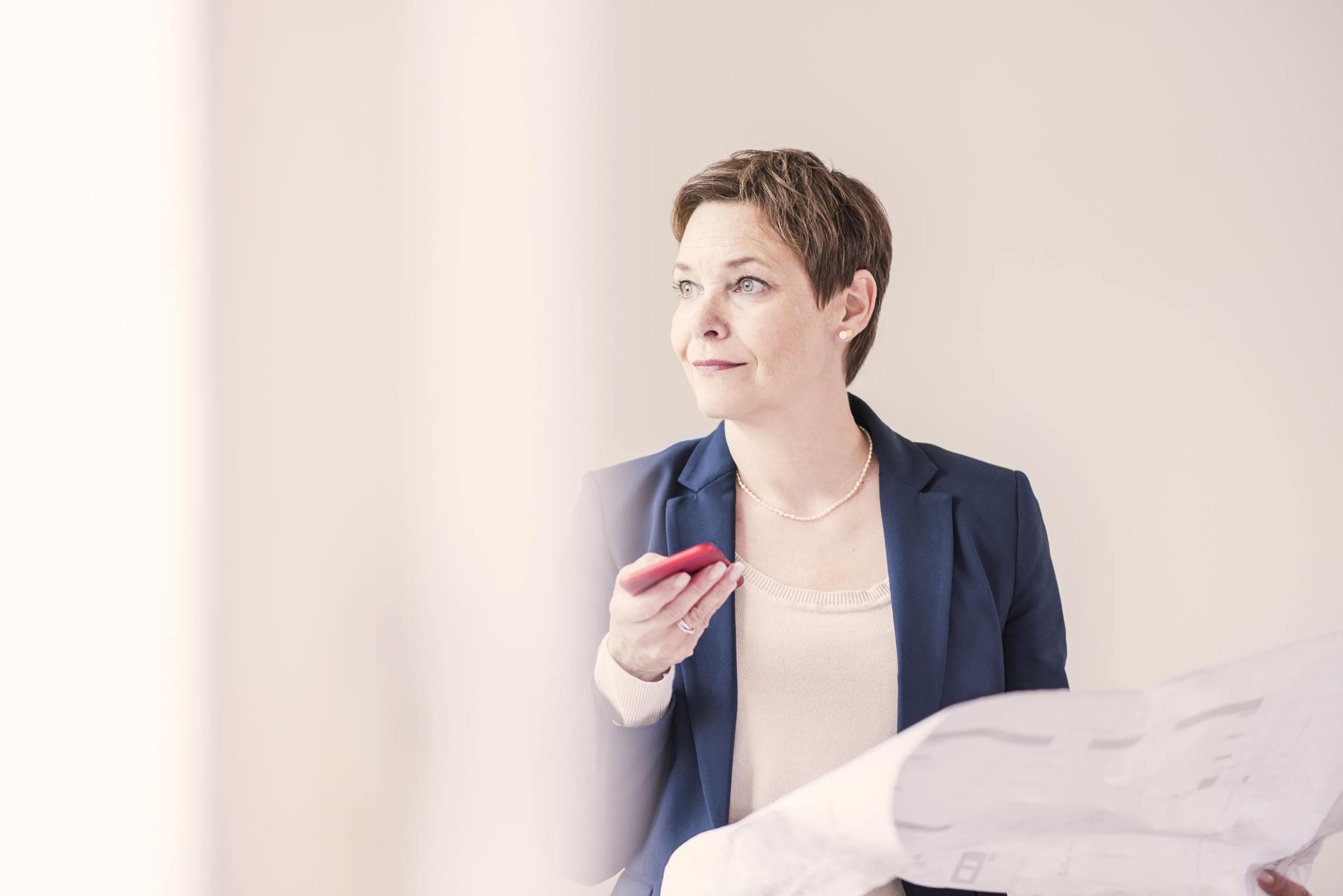 Eine Frau im Businessanzug hält in einer Hand ein Mobiltelefon mit einer roten Handyhülle und in der anderen ein großen Zettel und schaut dabei seitlich in die Ferne.