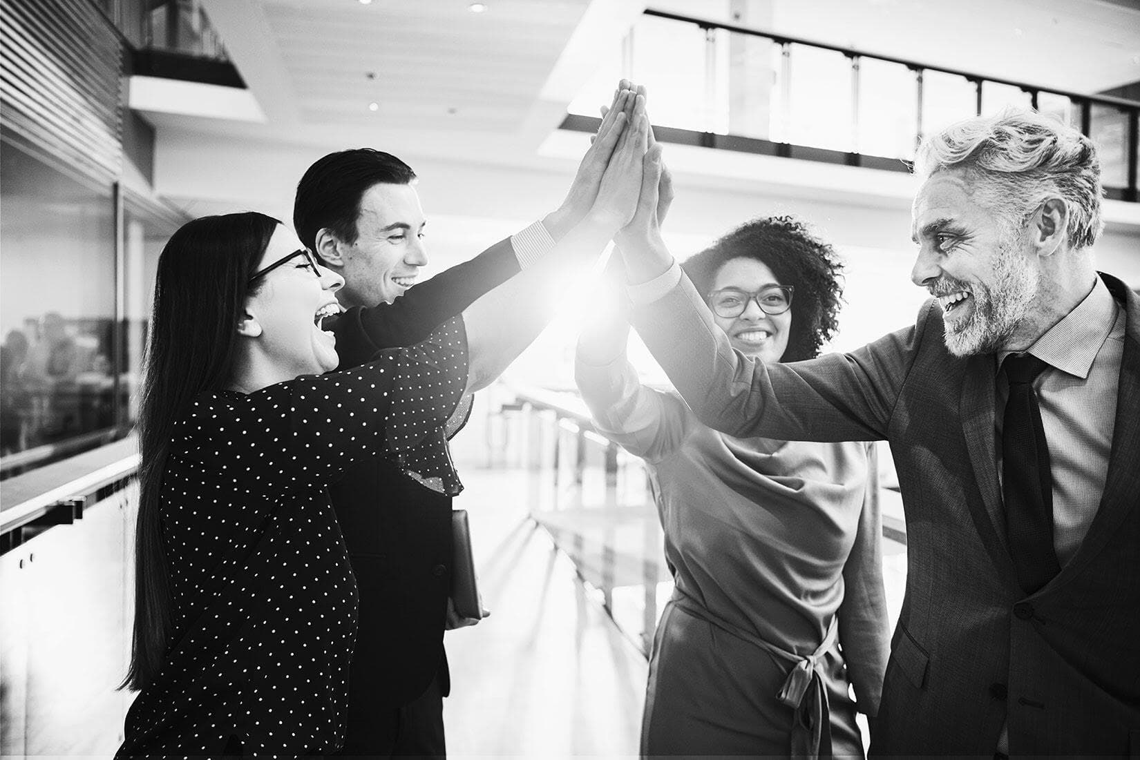 Vier Personen stehen in einer Reihe und geben sich gegenseitig High-Fives. Die Gesichter sind unscharf, im Hintergrund moderne Architektur mit großen Fenstern und einem Geländer.