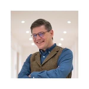 Eine Person mit unscharfem Gesicht steht mit verschränkten Armen. Die Person trägt ein blaues Hemd und eine braune Weste. Der Hintergrund scheint ein Innenraum mit weißen Wänden und Deckenleuchten zu sein.