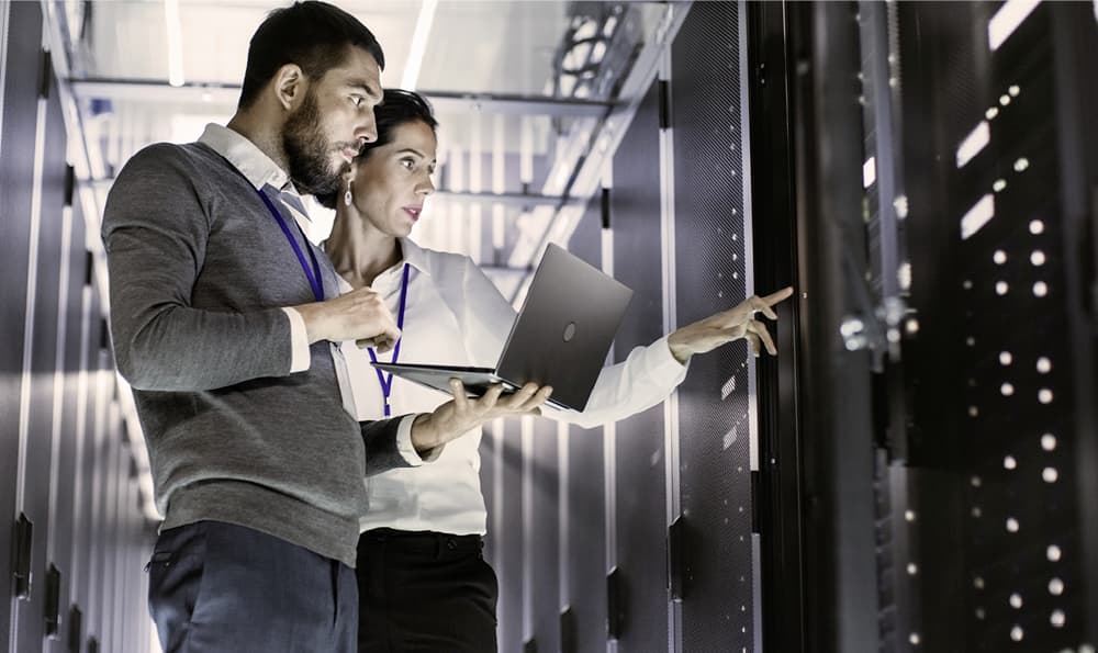 Zwei Personen in einem Serverraum, eine Person hält einen Laptop und die andere zeigt auf ein Server-Rack. Beide tragen ID-Karten um den Hals.