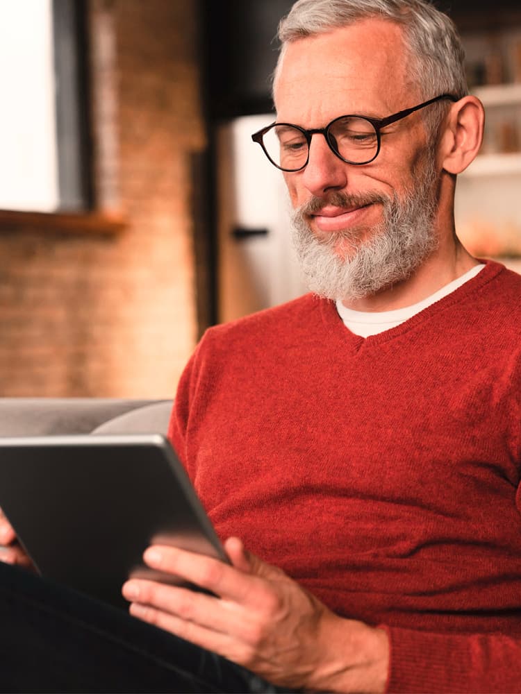 Eine ältere Person mit grauen Haaren und Bart, trägt eine Brille und einen roten Pullover, sitzt auf einem grauen Sofa und hält ein Tablet.