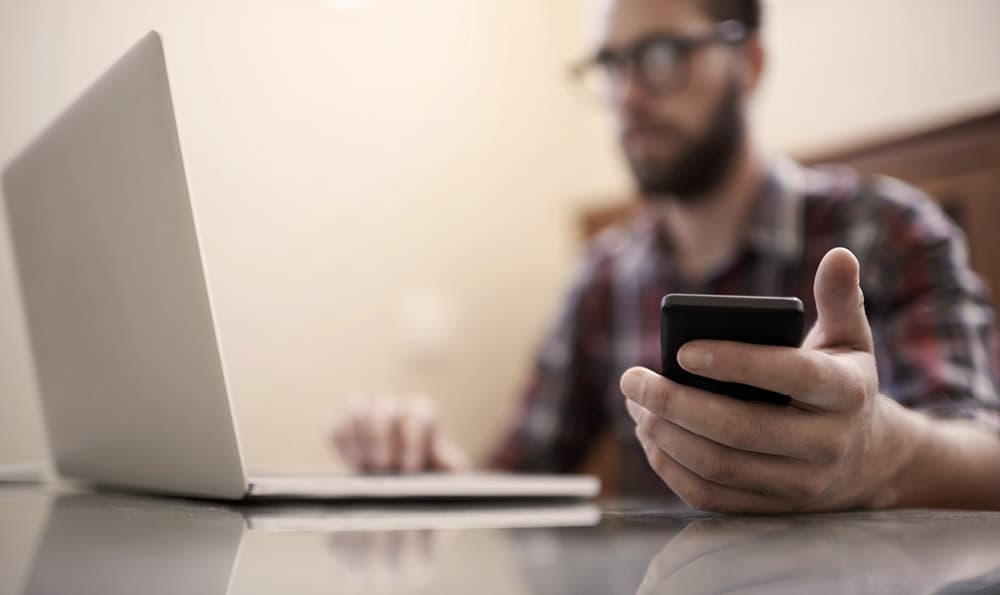 Eine Person sitzt an einem Schreibtisch, benutzt einen Laptop und hält ein Smartphone. Das Gesicht der Person ist unscharf, und der Fokus liegt auf den verwendeten Geräten.