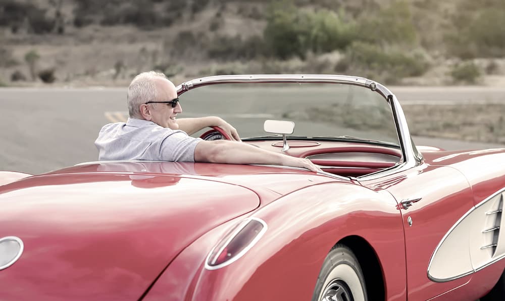 Eine Person fährt ein klassisches rotes Cabrio auf einer offenen Straße, aund das Auto hat ein elegantes Design mit Chromdetails und weißen Akzenten.