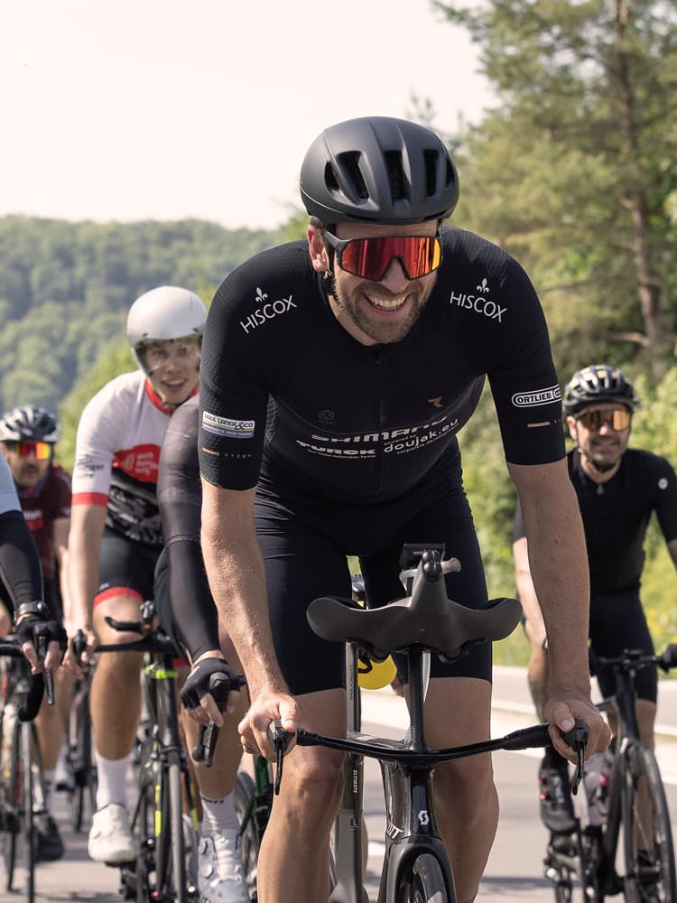 Eine Gruppe von Radfahrern fährt auf einer Straße; im Vordergrund ein Fahrer in schwarzer Kleidung mit „HISCOX“-Logo auf den Ärmeln, im Hintergrund weitere Radfahrer und grüne Bäume entlang der Strecke.