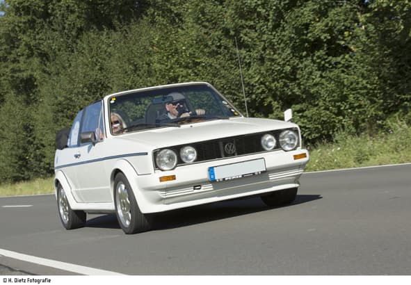 Weißes Cabriolet mit zwei Personen fährt auf einer Straße, im Hintergrund Bäume; blaues Nummernschild sichtbar.