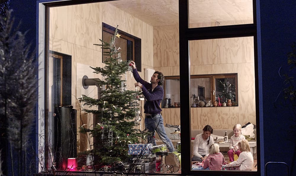 Eine Familie schmückt im Haus einen Weihnachtsbaum. Eine Person steht auf einem Stuhl und hängt eine Kugel auf, während drei weitere am Tisch sitzen. Draußen ist es dunkel.