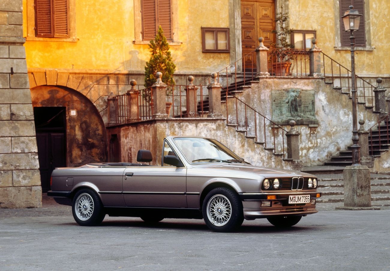 Ein grauer BMW 325i Cabrio E30 parkt in einer Gasse.