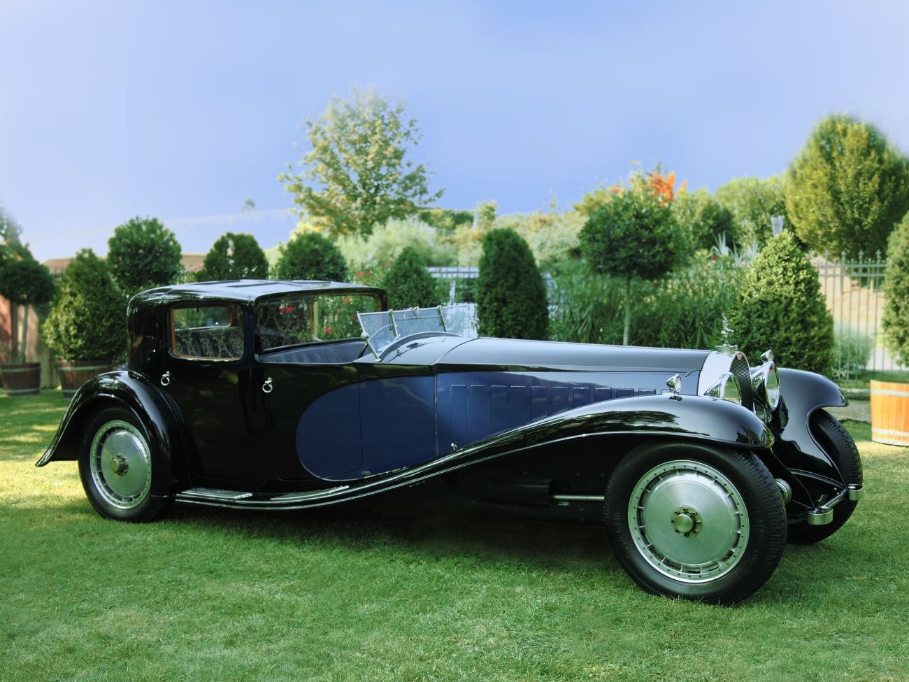 Der dunkelblaue Bugatti Royale 1926 parkt auf einer grünen Wiese vor blühenden Bäumen.