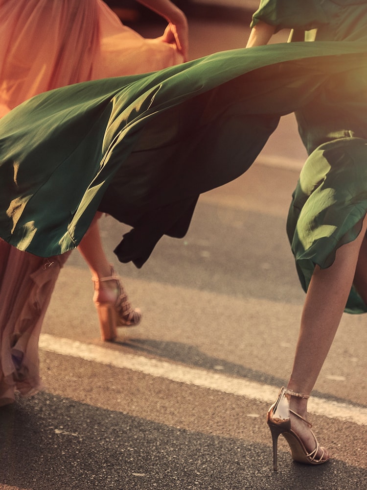 Zwei Frauen laufen im Sonnenlicht über eine Straße und ihre langen Kleider wehen im Wind