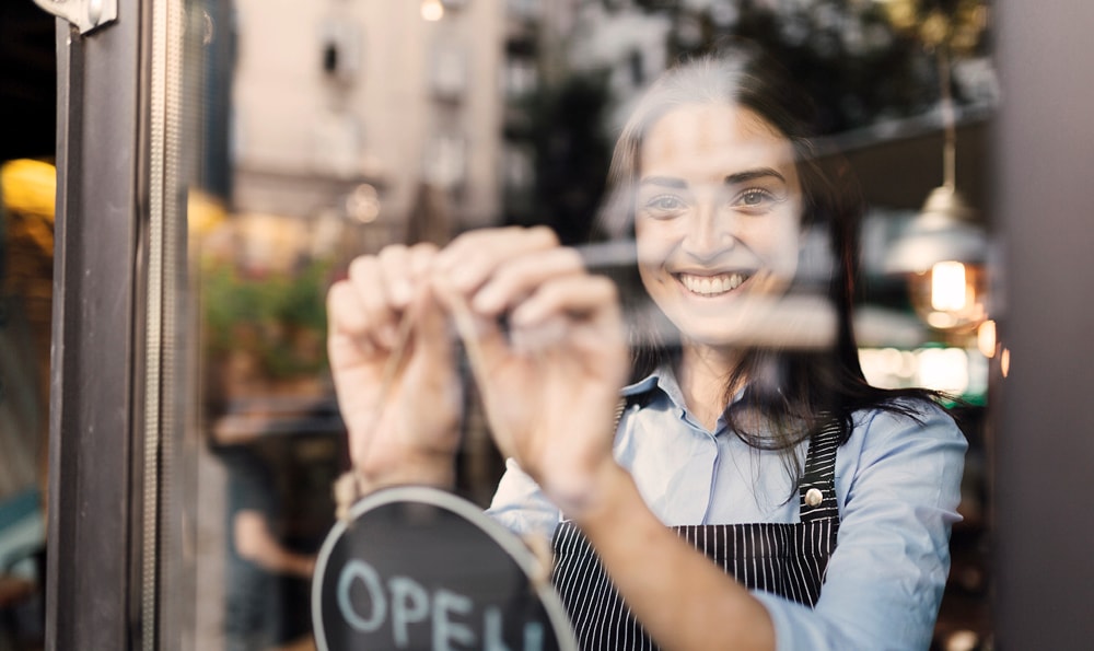Eine lächelnde Frau in einer gestreiften Schürze öffnet ein Café und richtet ein Schild an der Glastür aus. Die Szene ist warm und einladend, mit verschwommener Straßenansicht draußen.