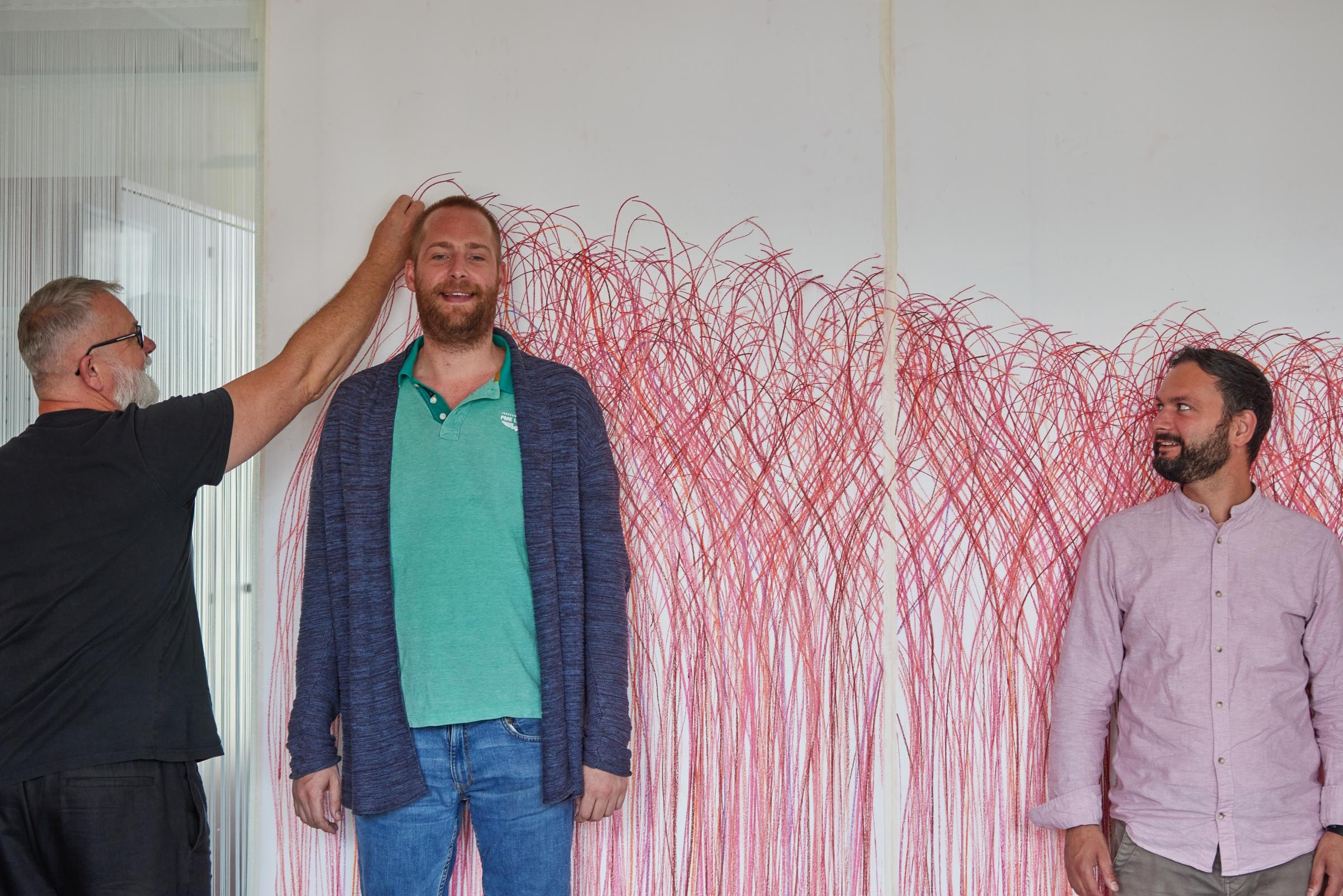 Drei Männer stehen vor einem mit roten Kreidefarben gemalten großen Wandbild, auf das die Umrisse des Mannes in der Mitte gezeichnet werden.