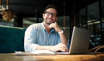 Ein junger Mann mit Brille und in einem blauen Hemd sitzt vor seinem Laptop und lächelt in die Kamera