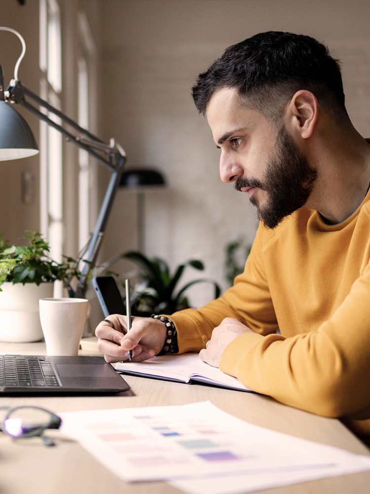 Ein junger Mann mit gelbem Pullover sitzt an einem Schreibtisch und arbeitet am Laptop