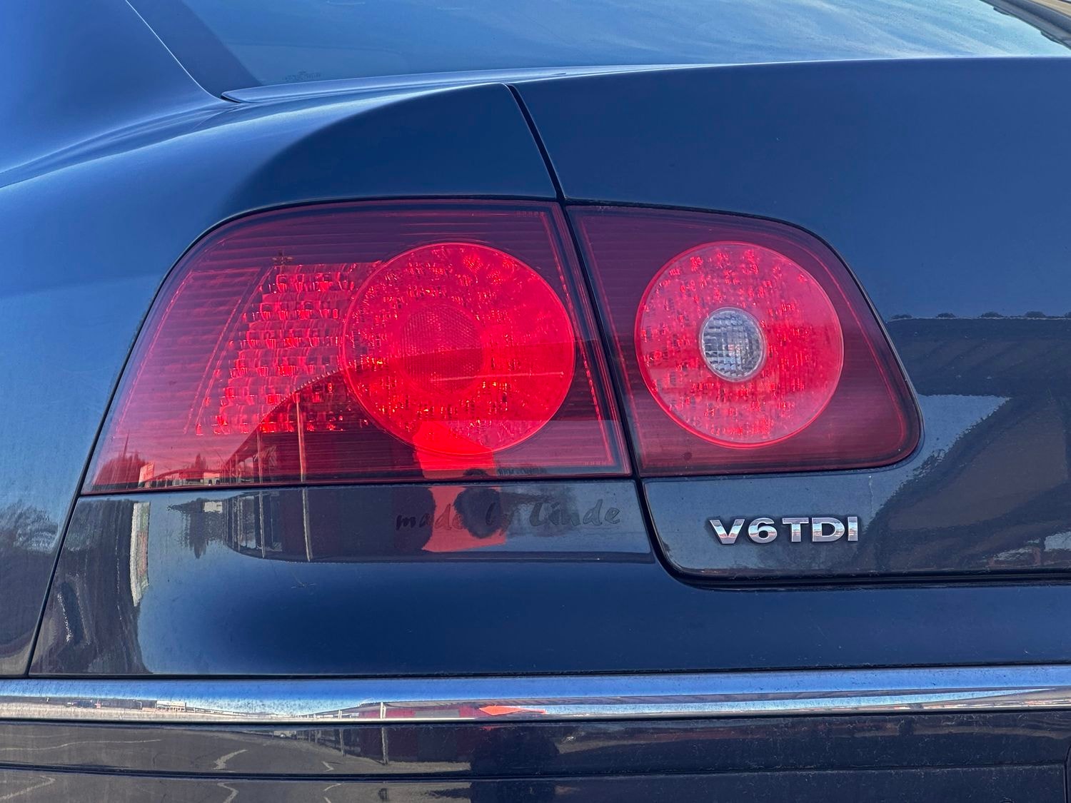Nahaufnahme des Hecks eines schwarzen Autos mit roten Rückleuchten und einer verchromten V6TDI-Plakette, die das Sonnenlicht reflektiert. Das Bild vermittelt einen eleganten und modernen Eindruck.