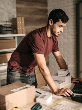 Junger Mann in rotem T-shirt steht an einem Tisch in einem Lager und bearbeitet Bestellungen