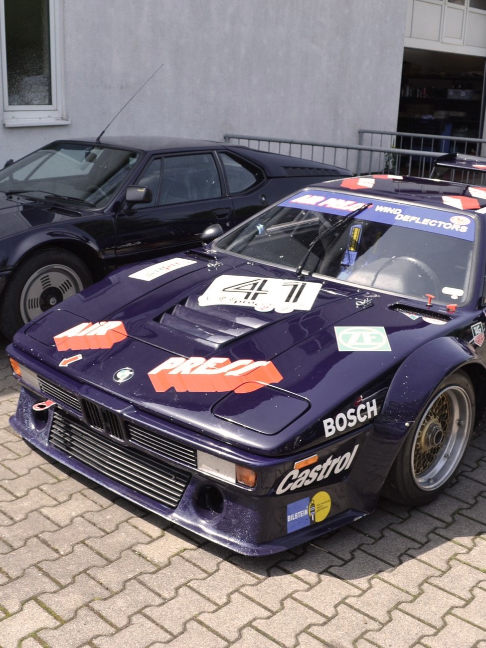 Auf einer Ziegelstein-Auffahrt sind zwei Oldtimer geparkt, davon ist einer der BMW M1.