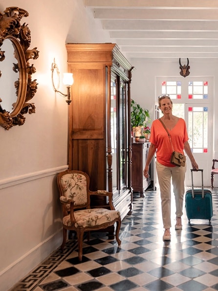 Eine lächelnde Frau in einem orangefarbenen Oberteil zieht einen blaugrünen Koffer durch einen hellen, eleganten Flur mit kariertem Boden, einem Vintage-Sessel und einem dekorativen Spiegel.