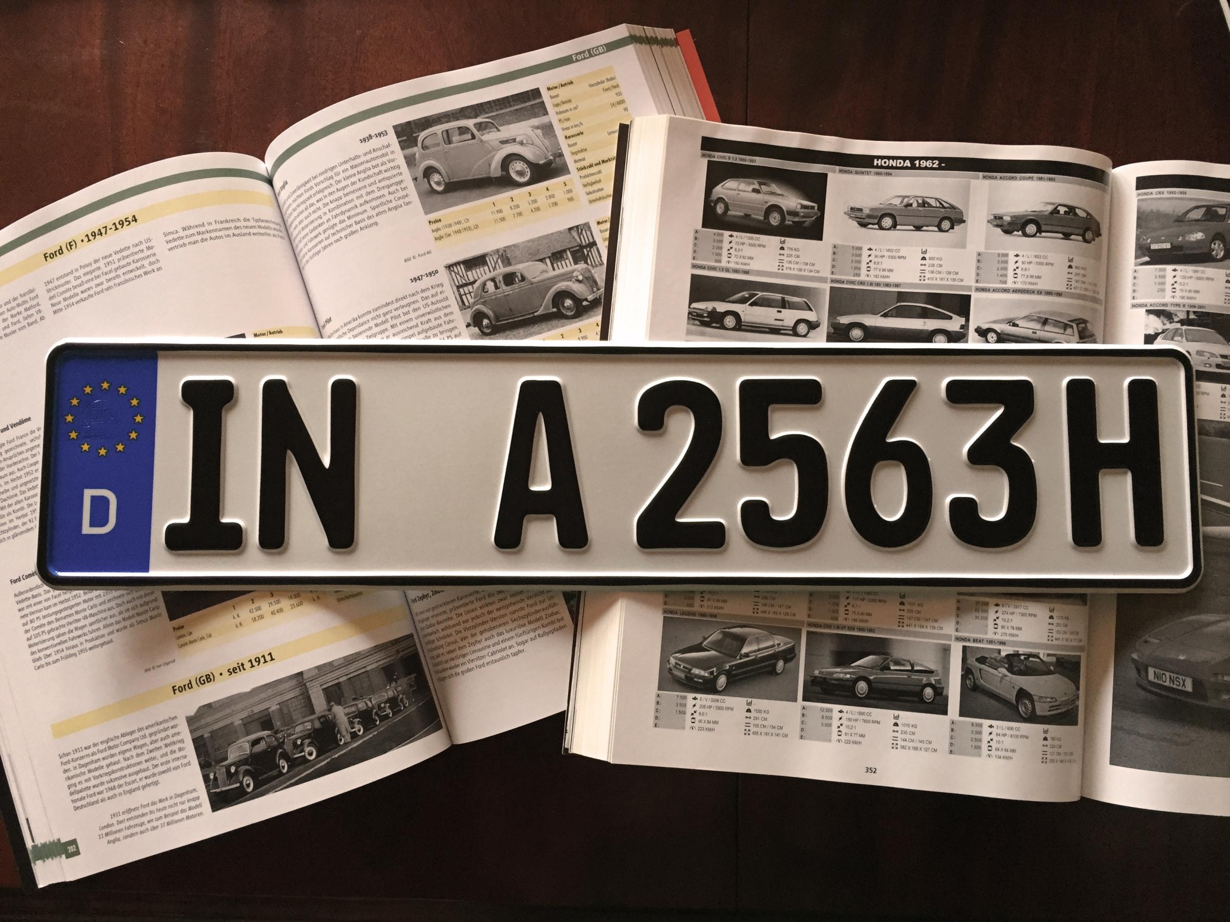Ein deutsches Autokennzeichen mit der Aufschrift 'IN A 2563H' liegt auf einem aufgeschlagenen Automagazin, das verschiedene Autos und deren Spezifikationen zeigt.