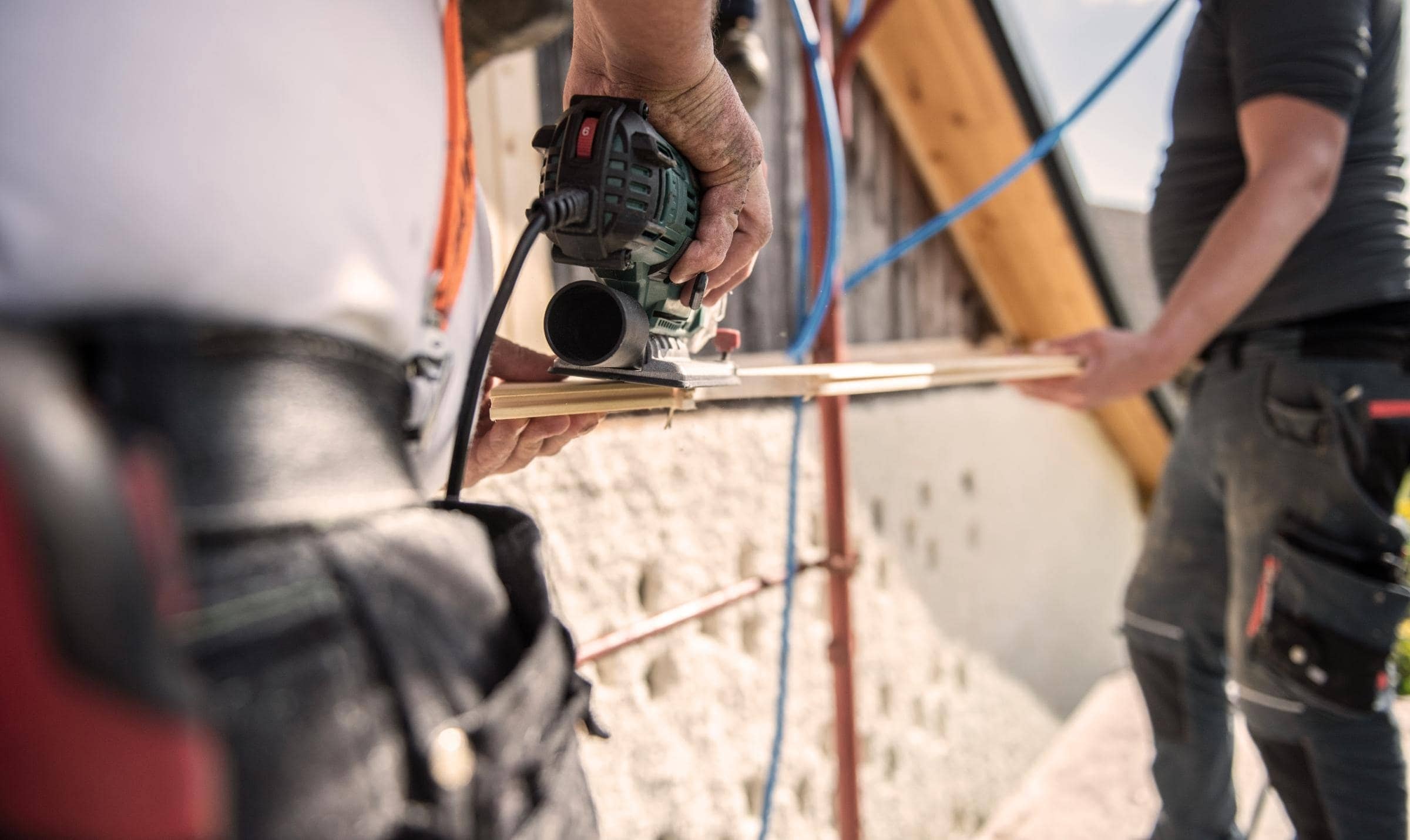 Zwei Handwerker in Arbeitskleidung arbeiten an einem Haus; einer schleift, während der andere ein Holzbrett hält.