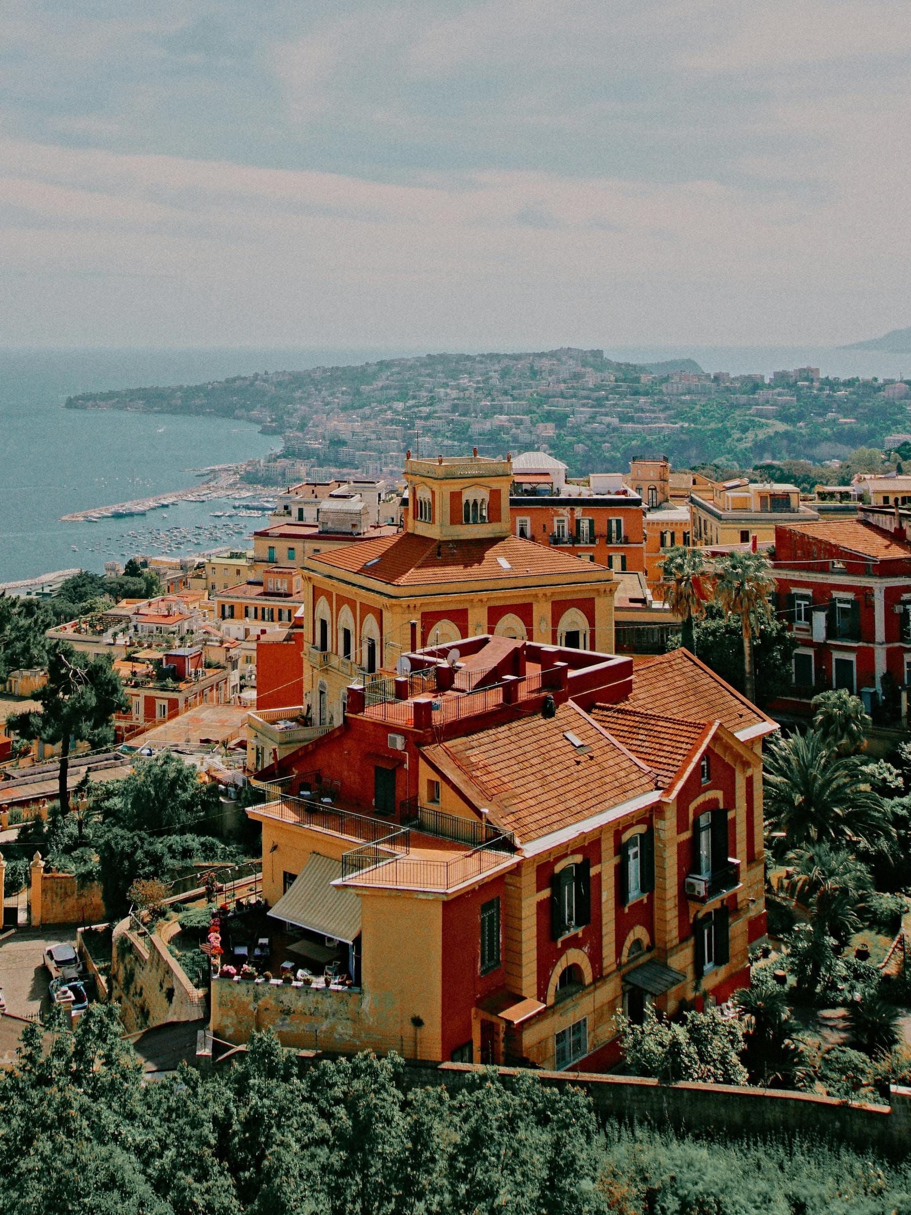 Im Mittelpunkt steht ein gelb rotes Haus auf einem bewachsenen Hügel in Italien. Im Hintergrund sieht man das Meer und eine Küstenlandschaft.