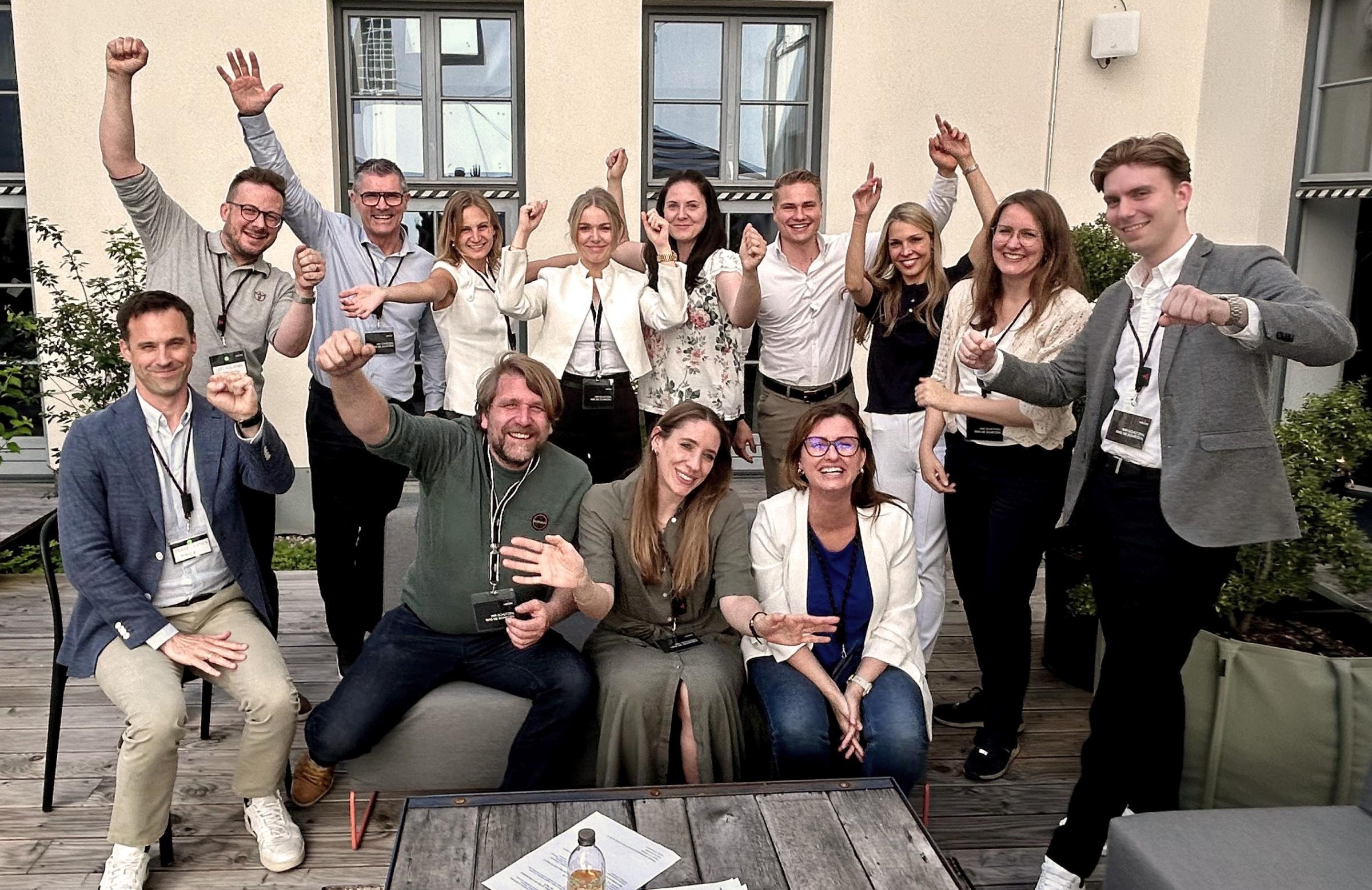 Eine Gruppe von fünfzehn Personen posiert auf einer Terrasse für ein Foto. Einige sitzen auf Sofas, während andere stehen. Alle haben ihre Gesichter verdeckt und tragen Namensschilder um den Hals. Die Stimmung scheint fröhlich und ausgelassen zu sein, da viele Personen ihre Arme in die Luft strecken oder Daumen hoch zeigen