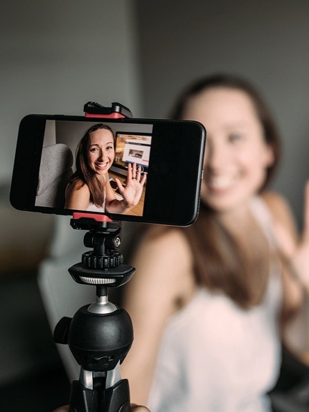 Lächelnde Influencerin sitzt vor ihrem PC und hält eine Vlogging-Kamera in der Hand und filmt sich selber