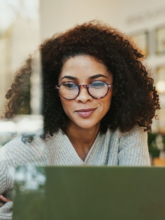 Eine Frau mit lockigem Haar und Brille sitzt vor einem Laptop und blickt lächelnd auf den Bildschirm.