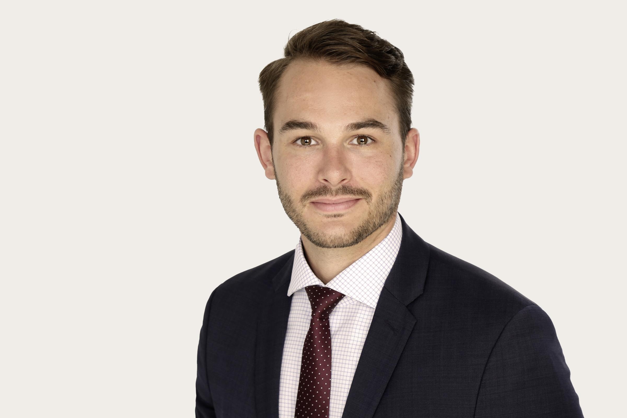 Moritz Assenmacher trägt ein dunkles Sakko, ein weißes Hemd mit Karomuster und eine rote Krawatte mit kleinen weißen Punkten.