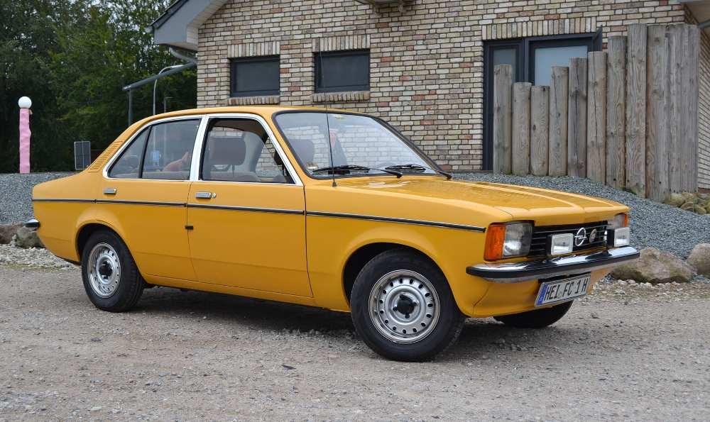 Gelbes Vintage-Auto auf einem Kiesplatz vor einem Backsteingebäude mit Holzzaun.