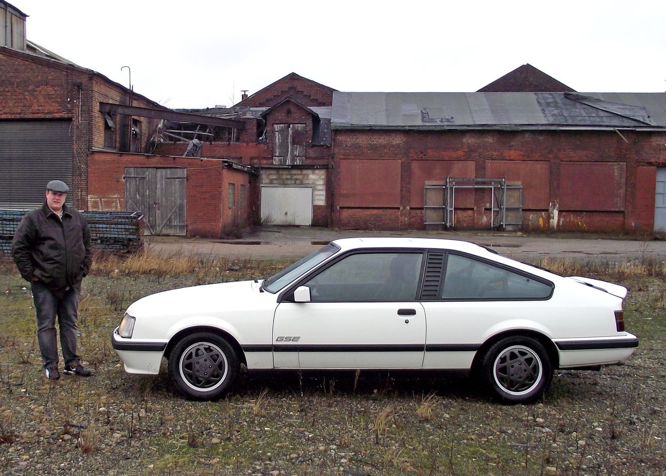 Weißer parkender Opel Monza GSE mit Seitenansicht vor einem Haus daneben steht ein Mann in Jacke und Hut.