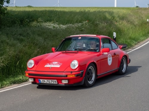 Ein leuchtend roter Porsche 911 im Rallye-Stil aus früheren Zeiten fährt an einem sonnigen Tag eine Landstraße entlang, hinter ihm erstrecken sich grüne Felder und Windräder.