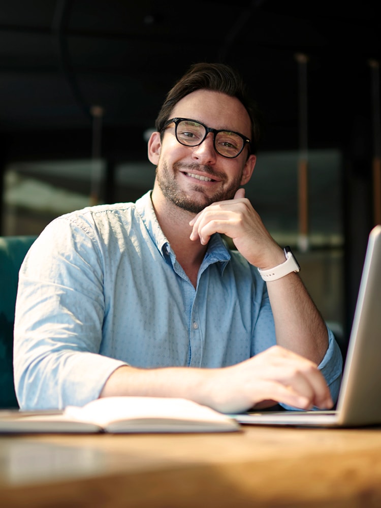 Ein junger Mann mit Brille und in einem blauen Hemd sitzt vor seinem Laptop und lächelt in die Kamera