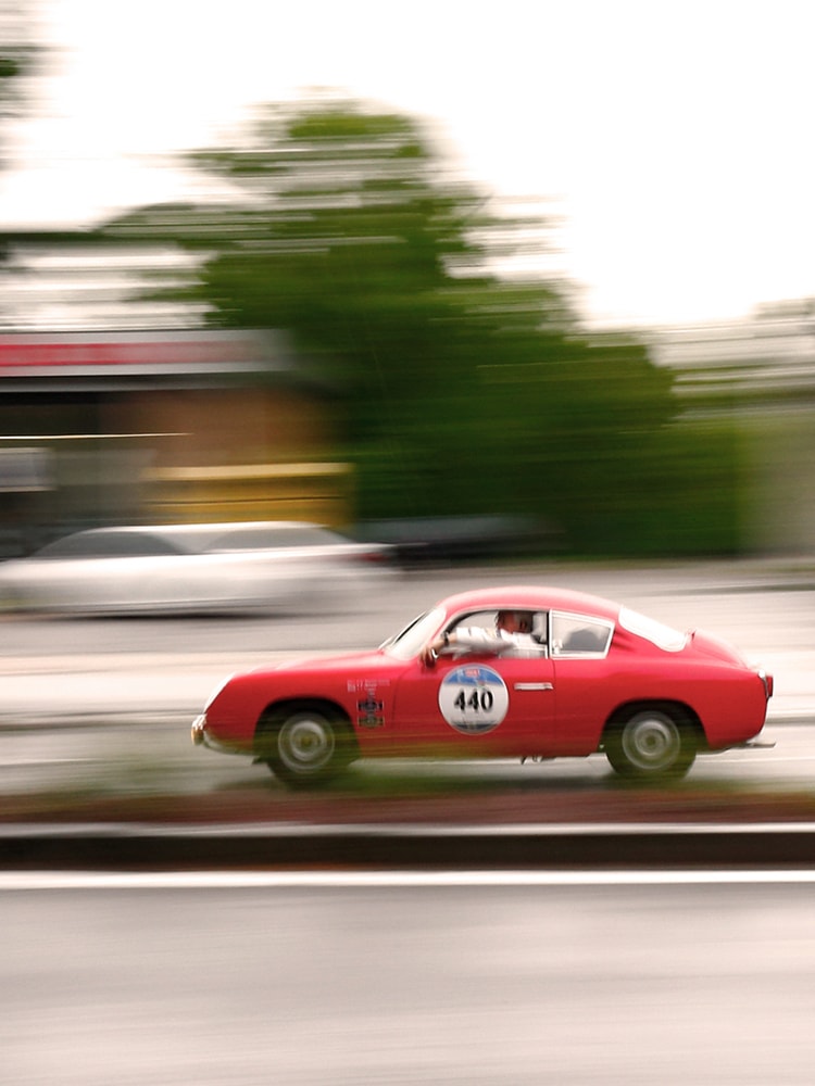 Roter Oldtimer-Rennwagen mit Startnummer fährt auf nasser Straße, Bewegungseffekt sichtbar.