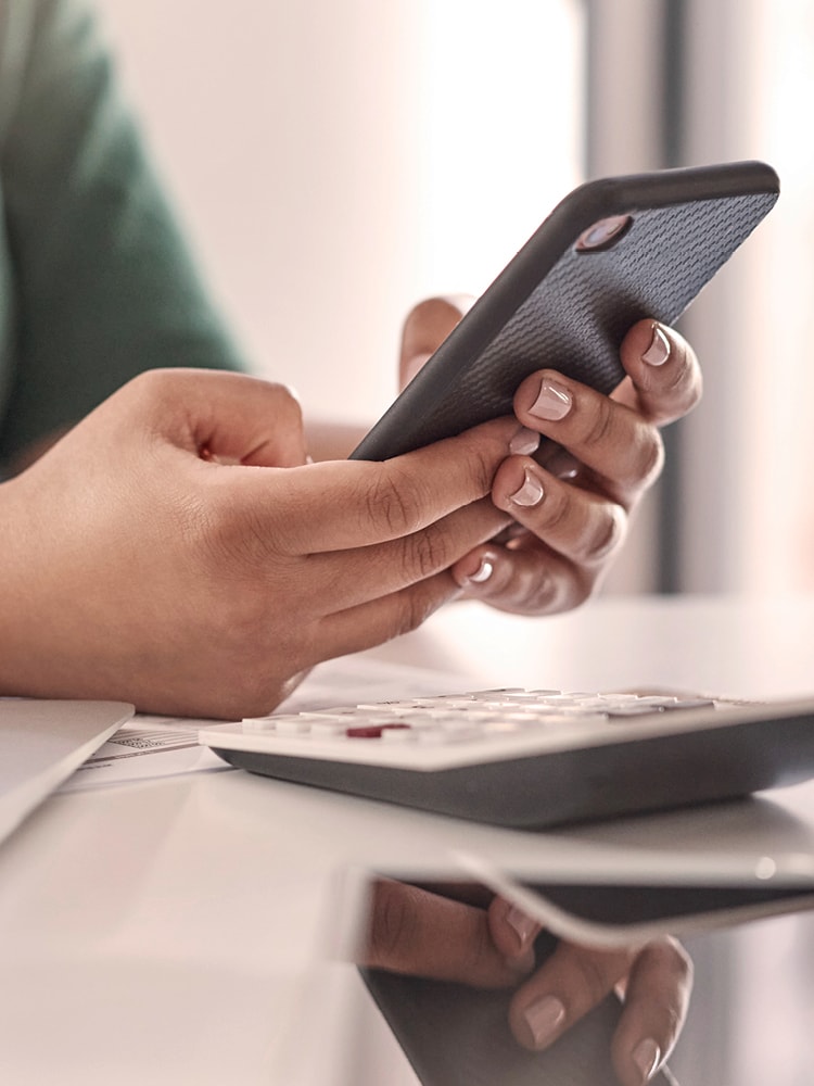 Jemand arbeitet am Smartphone, daneben stehen ein Laptop und ein Taschenrechner