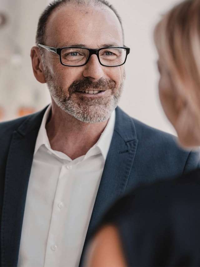 Ein Mann in einem dunklen Anzug und weißem Hemd spricht mit einer Frau.
