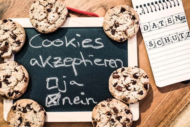 Eine kleine Tafel, umgeben von Schokoladenkeksen, mit der Aufschrift "Cookies akzeptieren". Darunter sind zwei Optionen: "ja" mit einem leeren Kontrollkästchen und "nein" mit einem angekreuzten Kontrollkästchen. Rechts von der Tafel liegt ein Notizblock mit dem Wort "DATENSCHUTZ"