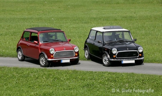Zwei Oldtimer-Minis, ein roter und ein schwarzer, parken nebeneinander auf einem Weg umgeben von einer grünen Wiese.