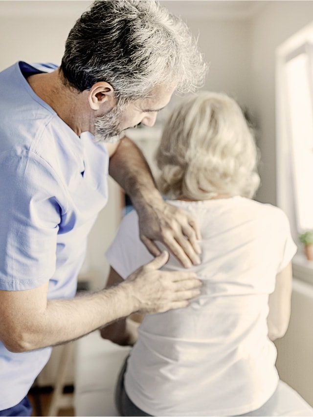 Ein Krankenpfleger in einem blauen Kittel untersucht den Rücken einer Frau mit weißen Haaren.