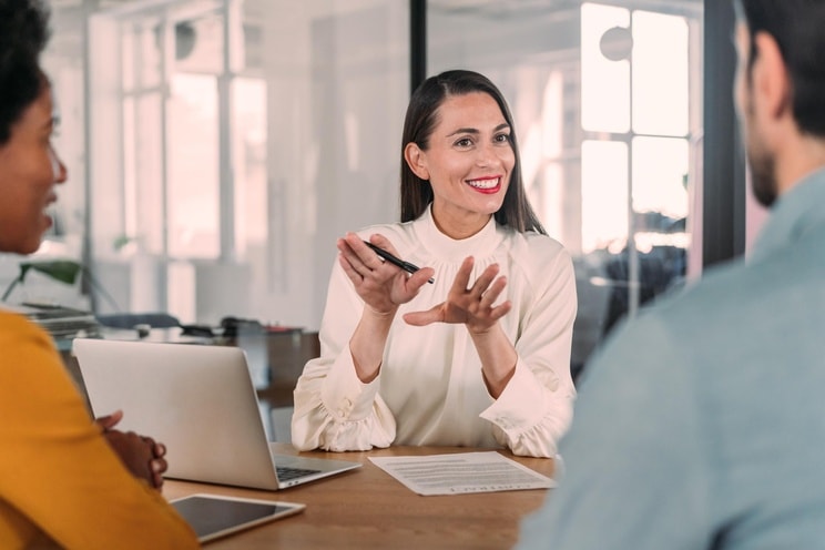 Beraterin in weißer Bluse und rotem Lippenstift sitzt vor einem Laptop und Papieren. Sie berät zwei Personen, die an einem Schreibtisch sitzen.