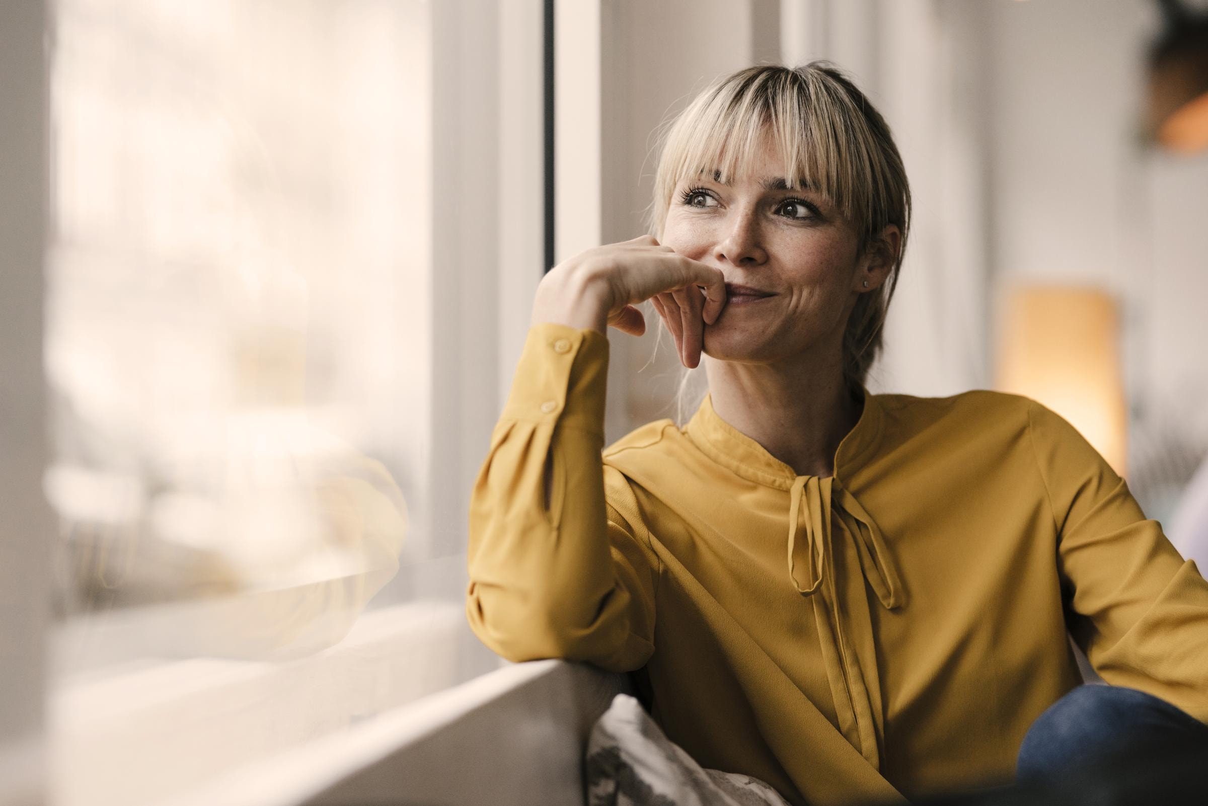 Eine Frau mit kurzen blonden Haaren und einem Pony sitzt vor einem Fenster und blickt lächelnd in die Ferne.