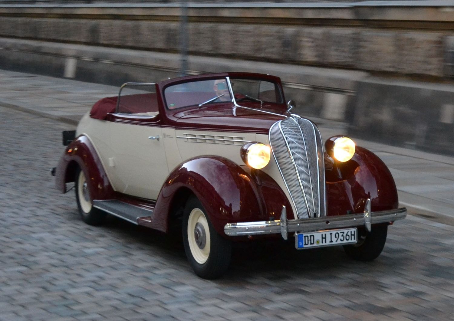 Ein klassisches rot-weißes Cabriolet aus den 1930er Jahren fährt auf einer gepflasterten Straße entlang einer historischen Steinmauer. Das Auto hat runde Scheinwerfer und ein markantes Chromgitter an der Front.