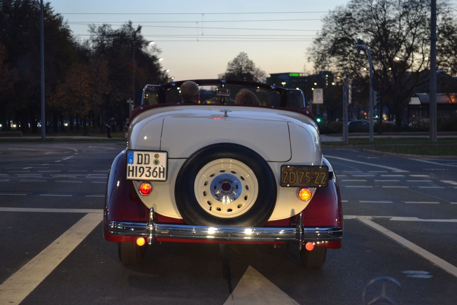 Oldtimer-Cabriolet fährt in der Dämmerung auf einer Straße.Die Rücklichter sind eingeschaltet, und im Hintergrund sind Bäume und Gebäude zu sehen.